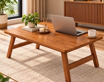 May include: A rectangular wooden coffee table with four angled legs, featuring a smooth, warm-toned finish. The table surface holds a silver laptop, a white ceramic mug on a small coaster, a small potted green plant, and a glass reed diffuser.