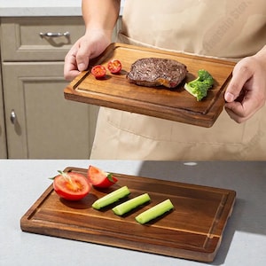 May include: A rectangular wooden cutting board with a groove around the edge. The board holds a cooked steak, cherry tomatoes, and broccoli. Another board shows tomatoes and celery. The boards are held by a person.