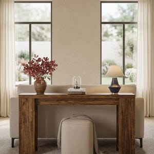 May include: A rectangular wooden console table with a dark brown wood grain finish, positioned behind a beige sofa. On the table sits a brown vase with red leaves, a glass cloche, and a dark blue table lamp with a beige fabric lampshade.
