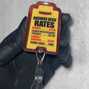 May include: A rectangular badge reel with a yellow face and red glitter border. The text reads: ANSWER DESK RATES, ANSWER $1.00, ANSWER WHICH REQUIRES THOUGHT $2.00, CORRECT ANSWERS $4.00, DUMB LOOKS FREE. It features a clear plastic strap and metal clip.