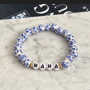 May include: White and blue floral bead bracelet with the word "MAMA" spelled out in white letter beads. Gold-colored beads separate the word from the floral beads. The bracelet is on a marble surface.