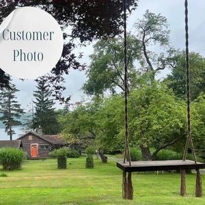 May include: A wooden swing hangs from ropes in a lush green yard. A small cabin with an orange door is visible in the background. The text "Customer Photo" is in the upper left corner.