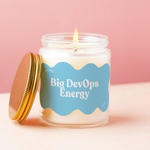 May include: A lit candle in a clear glass jar with a gold-colored metal lid placed to the side. The jar features a light blue, wavy-edged label with the white text Big DevOps Energy printed in a bold, rounded font on the front surface.