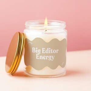 May include: A clear glass candle jar with a lit wick and a gold-colored metal lid placed to the side. The jar features a beige, wavy-edged label with the white text "Big Editor Energy" printed in a bold, serif font on the front surface.