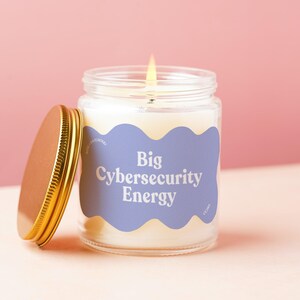 May include: A clear glass candle jar with a lit wick and a gold-colored metal lid resting beside it. The jar features a wavy-edged light purple label with the white text Big Cybersecurity Energy printed in a bold, serif font style.