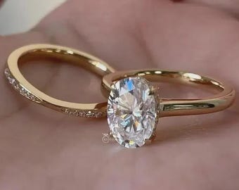 May include: A gold engagement ring with an oval-cut diamond and a matching gold band with small accent stones. The rings are set against a soft, neutral background, highlighting the sparkle of the diamond.