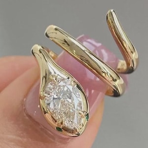 Può includere: Un anello aperto in oro con una pietra preziosa trasparente taglio marquise incastonata nella testa di un design a forma di serpente. La testa del serpente è accentuata da due piccole pietre verdi come occhi e la fascia in oro lucido si avvolge.