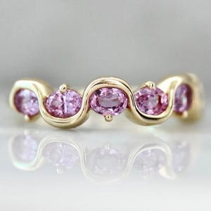 Peut inclure: Une bague en or dotée d'un anneau ondulé et sinueux serti de cinq pierres précieuses roses de taille ovale. La conception intègre une structure métallique fluide et continue qui entoure chaque pierre, créant un aspect équilibré et symétrique.