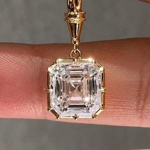 Puede incluir: Un colgante de oro con una gran piedra preciosa transparente de corte cuadrado y superficie facetada. La piedra está engastada en un marco de oro con garras en cada esquina, unida a una anilla de oro, suspendida por un hilo transparente.