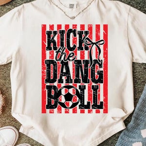 May include: Cream-colored t-shirt with a distressed graphic. The design features the text "KICK the DANG BALL" in black, set against a red and white striped background, with a soccer ball graphic. A black bow is also included.