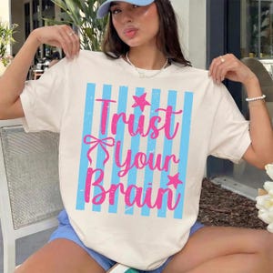Op de afbeelding: Crèmekleurig T-shirt met een blauwe en roze afbeelding. De afbeelding toont verticale blauwe strepen en de tekst "Trust Your Brain" in roze script, met ster- en strikaccenten. Een casual T-shirt voor elke dag.
