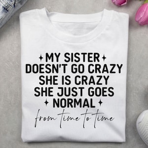 Pode incluir: T-shirt branco dobrado com texto preto: "MY SISTER DOESN'T GO CRAZY SHE IS CRAZY SHE JUST GOES NORMAL from time to time". A camisa parece ser de algodão ou tecido semelhante.
