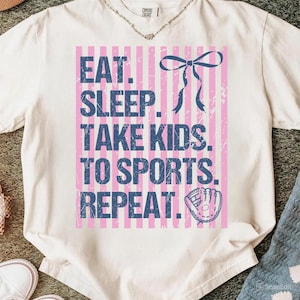 May include: Cream-colored t-shirt with a pink and blue graphic. The graphic reads "EAT. SLEEP. TAKE KIDS. TO SPORTS. REPEAT." in blue text over pink stripes. A blue bow and baseball glove are also featured.