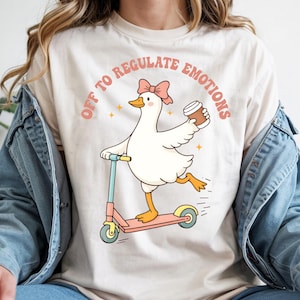 Puede incluir: Camiseta color crema con un dibujo de un ganso en un patinete rosa, sosteniendo una taza de café. El ganso lleva un lazo rosa y el texto "OFF TO REGULATE EMOTIONS" está arqueado encima en letras rosas. El diseño está impreso en una camiseta color crema.