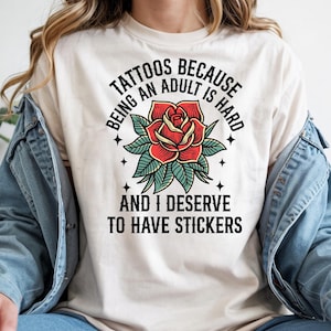 May include: Cream-colored t-shirt featuring a red rose tattoo design with green leaves. The text reads "Tattoos because being an adult is hard and I deserve to have stickers."