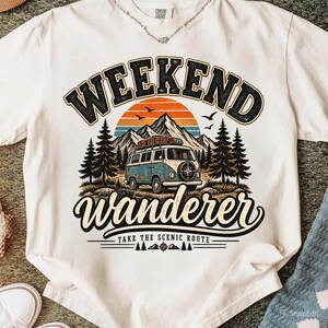 May include: A white t-shirt featuring a graphic print of a blue and white camper van parked in front of mountains and pine trees. The text reads WEEKEND WANDERER and TAKE THE SCENIC ROUTE, accompanied by a sunset illustration and flying birds.