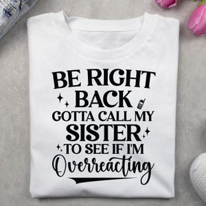 May include: White t-shirt with the text "BE RIGHT BACK GOTTA CALL MY SISTER TO SEE IF I'M Overreacting" in black. The design includes a phone graphic and star accents. The shirt is folded neatly.