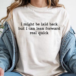 May include: Cream-colored t-shirt with the text "i might be laid back but i can lean forward real quick" in black. The shirt is a crew neck style and appears to be made of a soft material. The shirt is worn with a denim jacket.