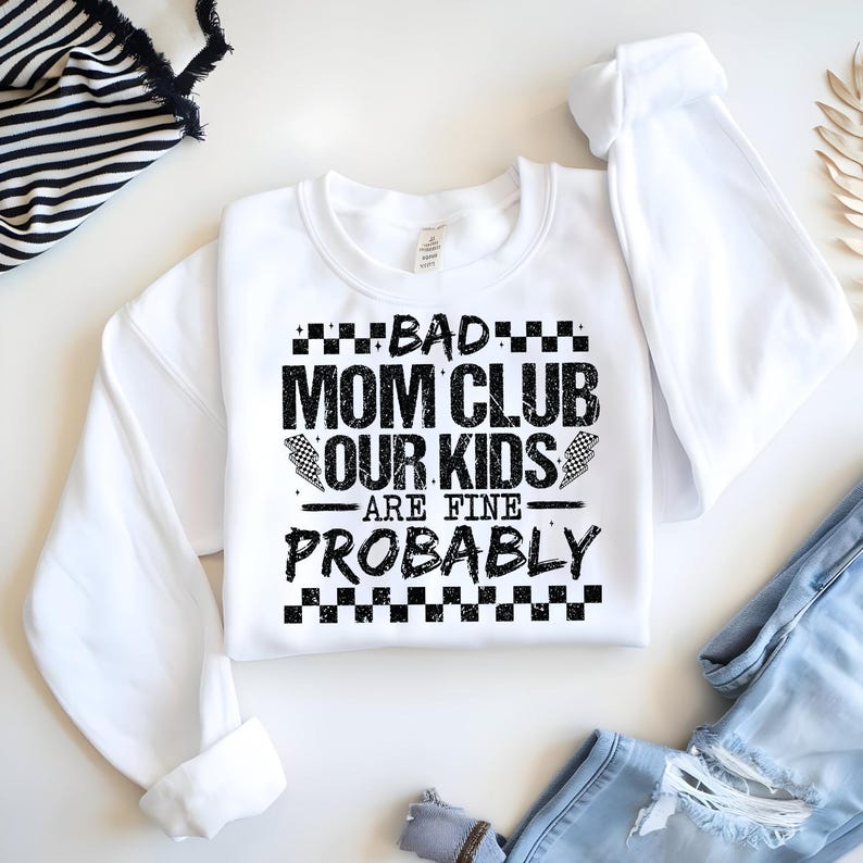 May include: White sweatshirt with the text "BAD MOM CLUB OUR KIDS ARE FINE PROBABLY" in black. The text is surrounded by a checkered pattern. The sweatshirt is folded with the sleeves showing.