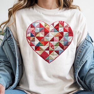 May include: Cream-colored t-shirt featuring a large, patchwork heart design. The heart is composed of various colorful fabric triangles in red, pink, blue, and green. The shirt has a classic crew neck and short sleeves.