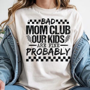 May include: Cream-colored t-shirt with the text "BAD MOM CLUB OUR KIDS ARE FINE PROBABLY" in black block letters. The design includes checkered flag graphics and a checkered border at the top and bottom of the text.