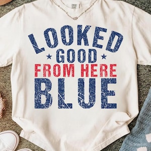 May include: Off-white t-shirt featuring the text "LOOKED GOOD FROM HERE BLUE" in distressed blue and red lettering. The shirt has a crew neck and short sleeves. A silver necklace is visible at the neckline.