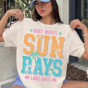 Op de afbeelding: Crèmekleurig T-shirt met de tekst "BOAT WAVES SUN RAYS LAKE DAYS" in oranje, blauw en groen. Het ontwerp bevat palmbomen en schelpgraphics, die een strandthema oproepen. Het is een casual, zomergericht kledingstuk.