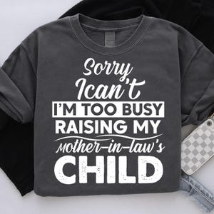 以下が含まれることがあります： 「Sorry, I can't, I'm too busy raising my mother-in-law's CHILD」の白文字がプリントされた濃いグレーのスウェットシャツ。カジュアルなスタイルで、普段使いに適しています。