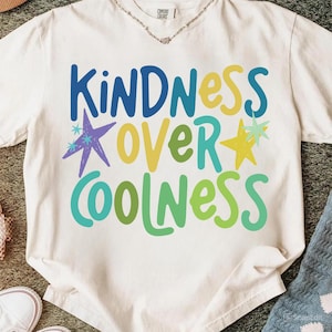 Może przedstawiać: Kremowa koszulka z napisem "KINDNESS OVER COOLNESS" w kolorze niebieskim, żółtym i zielonym, z gwiazdkami. Koszulka wykonana jest z miękkiego materiału i nadaje się do codziennego użytku.