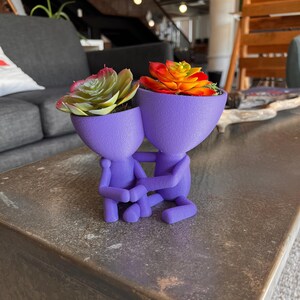 Puede incluir: Dos macetas moradas con forma humanoide tomadas de la mano, cada una con una planta suculenta. Una suculenta es verde y amarilla, la otra es naranja y roja. Las macetas están sobre una superficie oscura y texturizada.