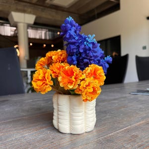 Peut inclure: Un arrangement floral avec des fleurs orange vif et bleues dans un vase crème matelassé. Les fleurs sont regroupées, créant une présentation pleine et colorée. Le vase présente un design texturé unique.