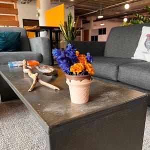 May include: A decorative ice cream cone-shaped vase filled with blue and orange flowers. The vase is light pink with a waffle cone texture and a brown chocolate-like top. The vase sits on a dark gray metal coffee table.