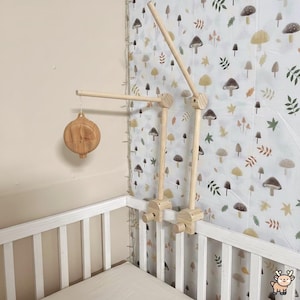 Puede incluir: Móvil de madera para bebé con un adorno redondo de madera colgante. El móvil está sujeto a una cuna blanca. El móvil tiene un acabado de madera natural. El fondo presenta un estampado de setas y hojas.