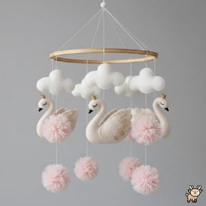 May include: A baby mobile featuring three white swans with gold crowns and accents, suspended below fluffy white clouds. Pink tulle pom-poms hang beneath the swans. The mobile is attached to a wooden hoop and hangs from a white string.