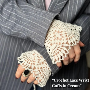 Puede incluir: Puños de muñeca de encaje de ganchillo color crema. Los puños se usan sobre las mangas de una blazer gris a rayas finas. Los puños tienen un diseño detallado y calado. El texto "Crochet Lace Wrist Cuffs in Cream" es visible.