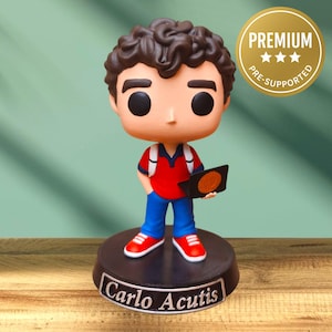 Puede incluir: Una figura de una persona con cabello rizado oscuro, vistiendo una camisa roja, pantalones azules y zapatos rojos. La figura sostiene un objeto negro con un círculo naranja. La base dice "Carlo Acutis". Una pegatina dorada dice "PREMIUM PRE-SUPPORTED".