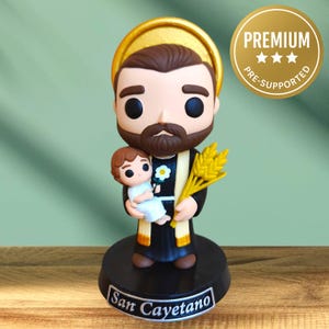 Puede incluir: Una figura de San Cayetano, sosteniendo a un niño y espigas de trigo. La figura tiene barba y cabello castaños, vistiendo una túnica negra con un halo dorado. La base es negra con el nombre "San Cayetano" en blanco. Una pegatina "PREMIUM" es visible.
