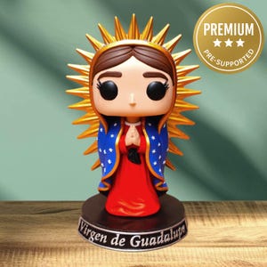 Puede incluir: Figura de la Virgen de Guadalupe, con un vestido rojo y una capa azul con estrellas blancas. La figura tiene un halo dorado y está sobre una base con el texto "Virgen de Guadalupe". Un círculo dorado con la palabra "PREMIUM" está en el fondo.