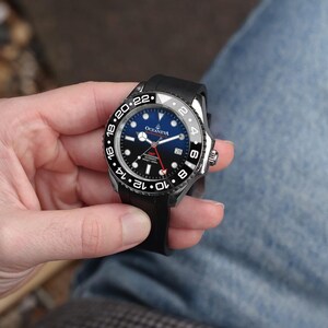 K&ouml;nnte beinhalten: Eine schwarze und silberne Armbanduhr mit blauem und schwarzem Zifferblatt wird in einer Hand gehalten. Die Uhr hat eine schwarze L&uuml;nette mit wei&szlig;en Ziffern und ein schwarzes Kautschukarmband. Das Zifferblatt zeigt die Marke "OCEANEVA".