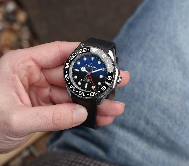 K&ouml;nnte beinhalten: Eine schwarze und silberne Armbanduhr mit blauem und schwarzem Zifferblatt wird in einer Hand gehalten. Die Uhr hat eine schwarze L&uuml;nette mit wei&szlig;en Ziffern und ein schwarzes Kautschukarmband. Das Zifferblatt zeigt die Marke "OCEANEVA".
