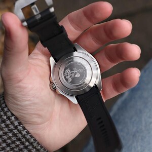 K&ouml;nnte beinhalten: Eine Edelstahl-Armbanduhr mit schwarzem Kautschukarmband wird in einer Hand gehalten. Das Zifferblatt der Uhr ist sichtbar, und die R&uuml;ckseite der Uhr weist ein eingraviertes Design auf. Es ist ein modernes Accessoire.