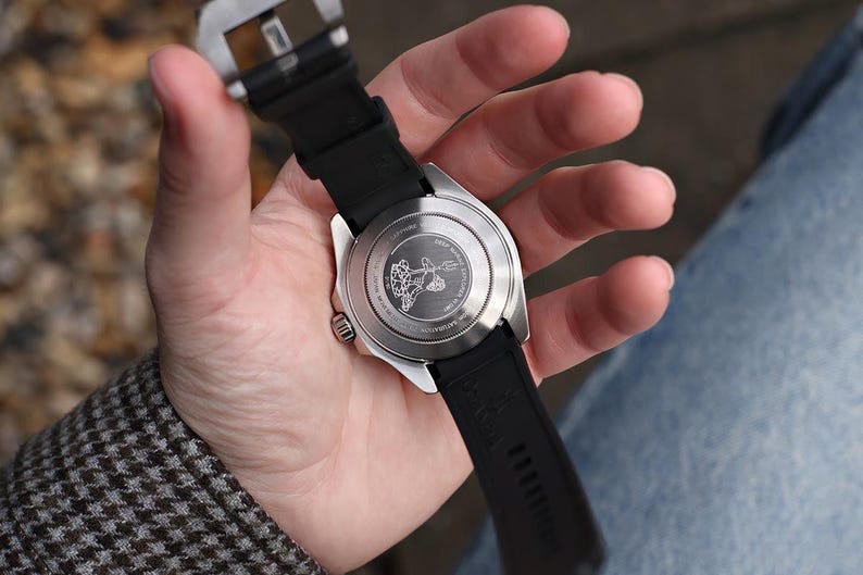 K&ouml;nnte beinhalten: Eine Edelstahl-Armbanduhr mit schwarzem Kautschukarmband wird in einer Hand gehalten. Das Zifferblatt der Uhr ist sichtbar, und die R&uuml;ckseite der Uhr weist ein eingraviertes Design auf. Es ist ein modernes Accessoire.
