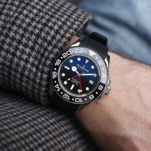 K&ouml;nnte beinhalten: Eine schwarze und silberne Armbanduhr mit blauem Zifferblatt und schwarzem Kautschukarmband. Die Uhr hat wei&szlig;e Ziffern und einen roten Sekundenzeiger. Die Uhr befindet sich am Handgelenk einer Person, mit einem Tweed-Jacken&auml;rmel im Hintergrund.