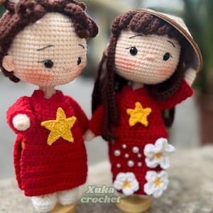 May include: Two handmade crochet dolls, a boy and a girl, holding hands. Both dolls wear red outfits with yellow star accents. The girl doll has long brown hair and a conical hat. The boy doll has short brown hair. The text "Xuka crochet" is visible.