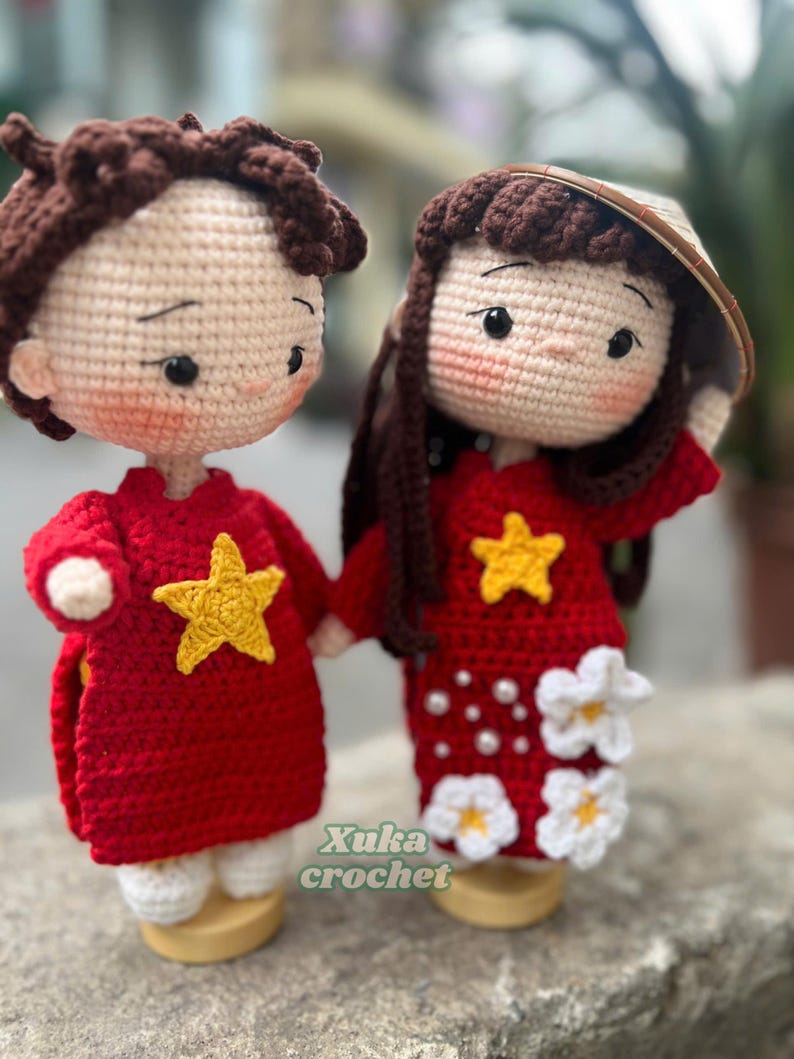 May include: Two handmade crochet dolls, a boy and a girl, holding hands. Both dolls wear red outfits with yellow star accents. The girl doll has long brown hair and a conical hat. The boy doll has short brown hair. The text "Xuka crochet" is visible.