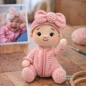 May include: A handmade crochet doll featuring a light peach skin tone, wearing a matching light pink onesie and a large, textured pink bow headband. The doll has embroidered black eyes, a small nose, and a red stitched mouth.