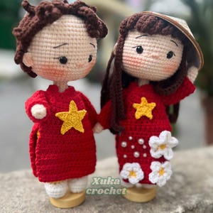 May include: Crocheted dolls of a couple, holding hands. They wear red outfits with yellow stars, and the woman has a conical hat. The dolls have brown hair and are on wooden bases. The text "Xuka crochet" is visible.