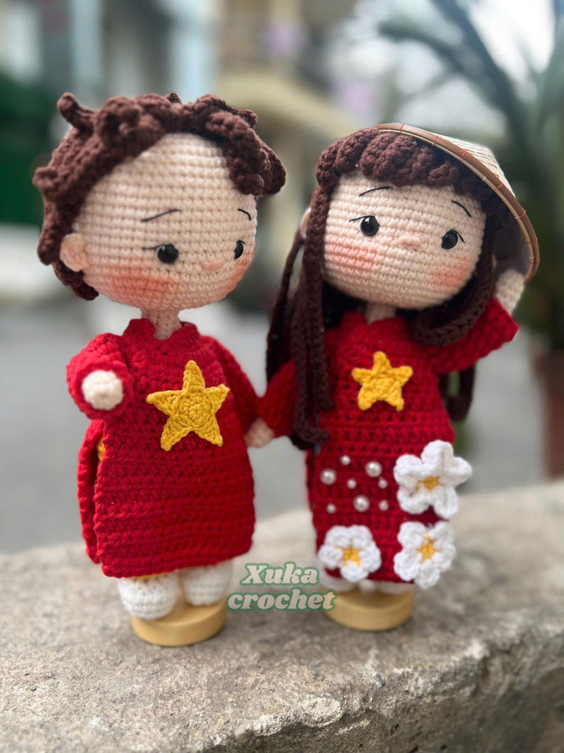 May include: Crocheted dolls of a couple, holding hands. They wear red outfits with yellow stars, and the woman has a conical hat. The dolls have brown hair and are on wooden bases. The text "Xuka crochet" is visible.