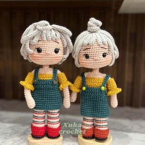 Puede incluir: Dos muñecas de ganchillo hechas a mano. Cada muñeca tiene el pelo gris, mejillas rosadas y lleva una camiseta amarilla y un peto verde azulado. Las muñecas están sobre bases de madera y llevan calcetines a rayas rojas y blancas. El texto "Xuka crochet" es visible.