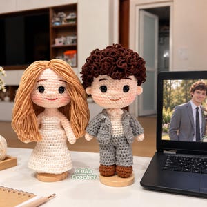 May include: Two crochet dolls standing on wooden bases. One doll has long light brown yarn hair and a white dress. The other doll has short dark brown curly yarn hair, a gray suit, and brown shoes. The text Xuka Crochet is visible between them.